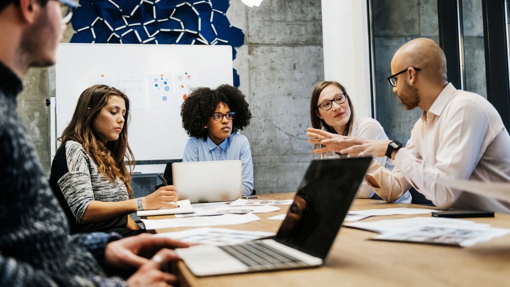 Image: A group of three young women and two men of different ethnicities hold a business meeting in a modern day office.