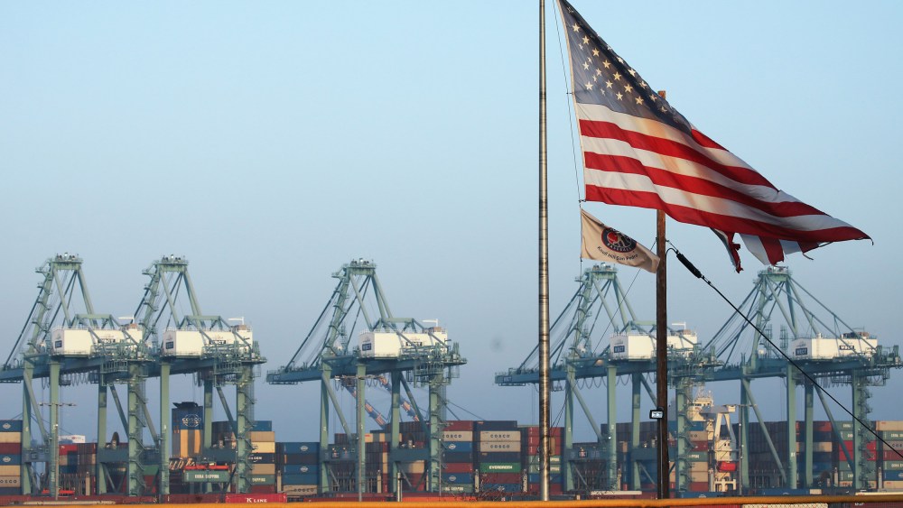 Image: An American flag flies with shipping containers stacked at the Port of Los Angeles in the background, which is the nation's busiest container port