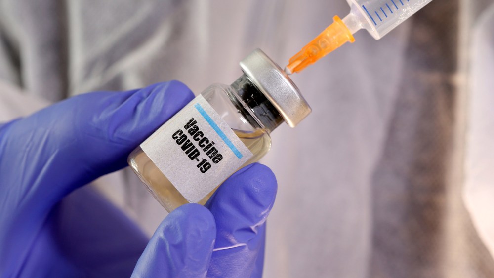 Image: A woman holds a small bottle labeled with a "Vaccine COVID-19" sticker and a medical syringe.