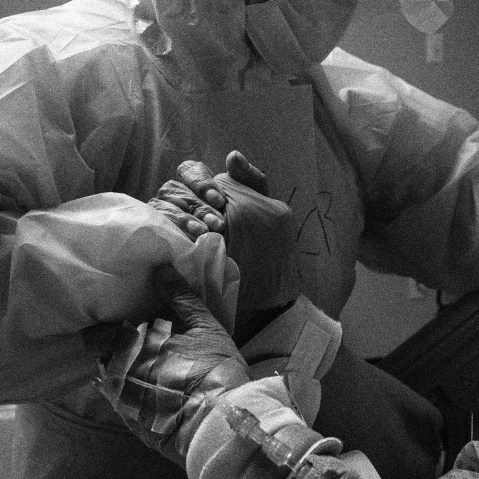 Image: A patient grips a doctor's hand in an emergency room. The doctor is wearing protective gear.