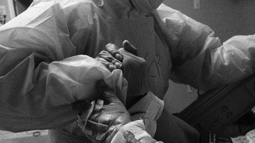 Image: A patient grips a doctor's hand in an emergency room. The doctor is wearing protective gear.