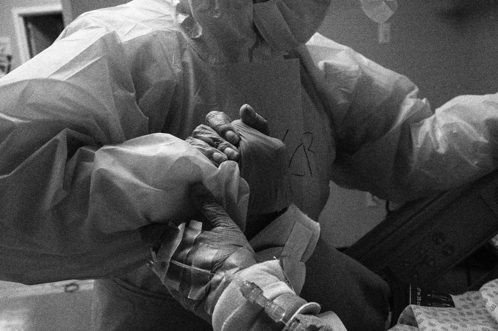 Image: A patient grips a doctor's hand in an emergency room. The doctor is wearing protective gear.