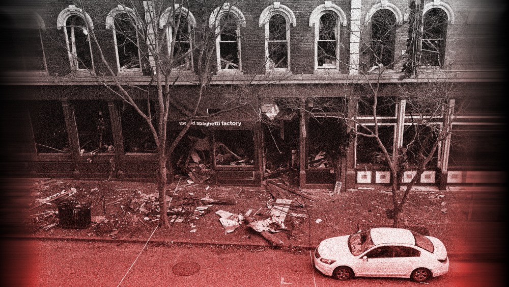 Image with a red overlay of debris remains on the sidewalk in front of buildings damaged in an explosion. A car is parked on the right.