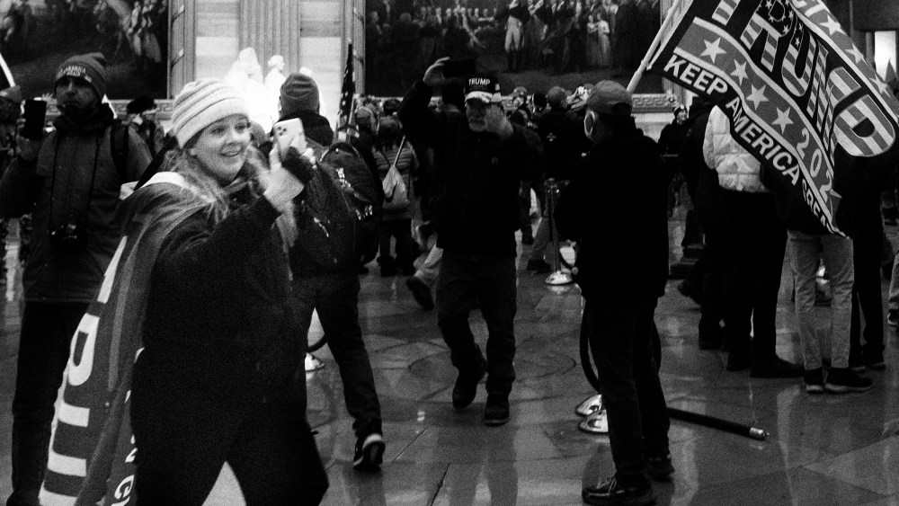 Image: Jenny Cudd wearing a cape that reads, "Trump" takes a photo with her phone amidst other rioters at the US Capitol's Rotunda.