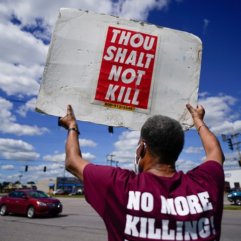 Image: Back of a person holding up a sign that reads,"Thou shalt not kill" and the back of their shirt reads,"No more killing".
