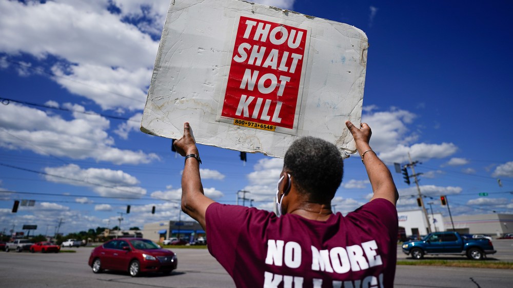 Image: Back of a person holding up a sign that reads,"Thou shalt not kill" and the back of their shirt reads,"No more killing".