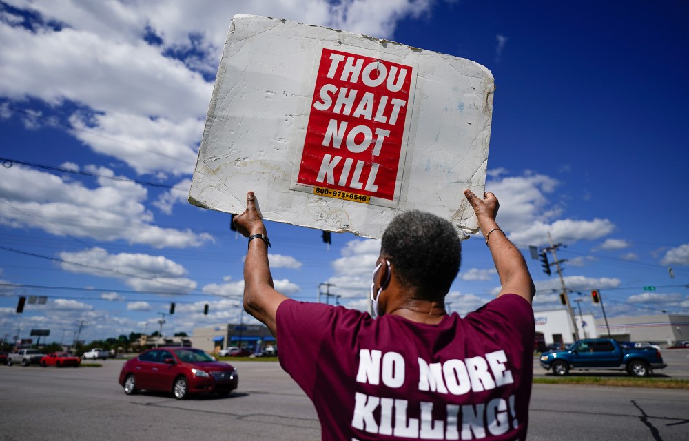 Image: Back of a person holding up a sign that reads,"Thou shalt not kill" and the back of their shirt reads,"No more killing".