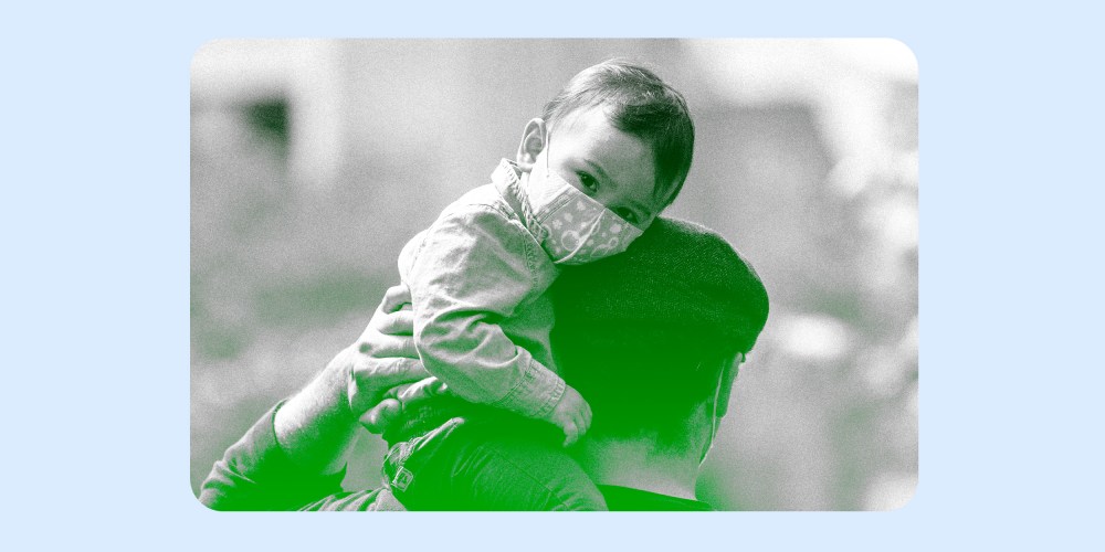Image: A young child wearing a mask sits on his fathers shoulders.