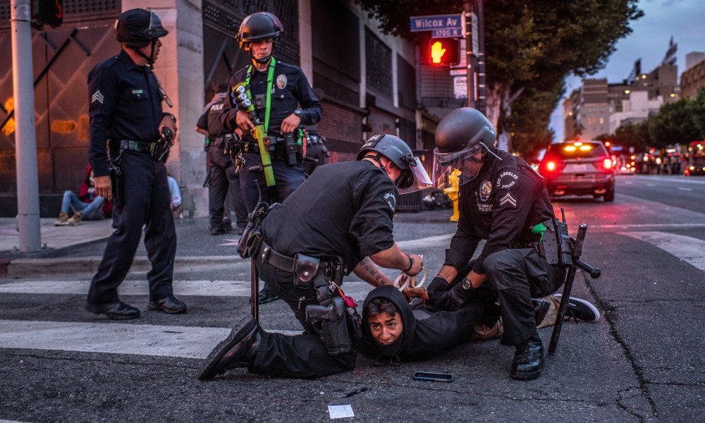 Image: Two police officers detain and handcuff a person on the street and two police officers stand by.