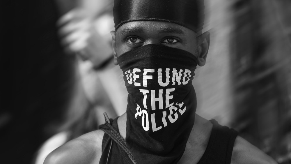 Image: A Black trans liberation activist wears a bandana that reads "Defund the Police" in New York City.