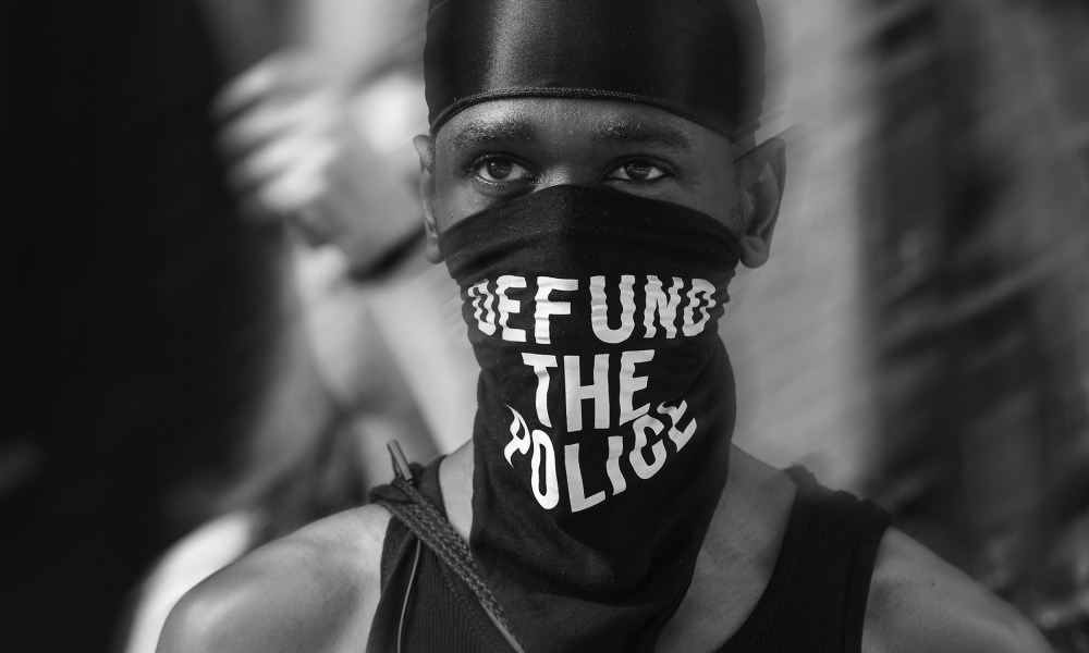 Image: A Black trans liberation activist wears a bandana that reads "Defund the Police" in New York City.
