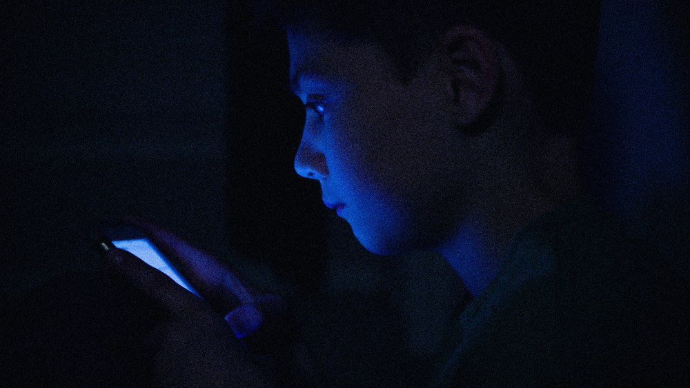 IMage: a teenage boy on his phone, face illuminated in the dark