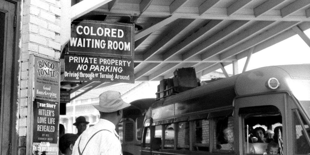 Image: People waiting at a bus station which has a sign that reads,"Colored Waiting Room".