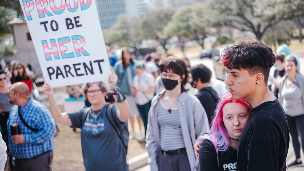 Image: A demonstrator holding a sign that reads,"Proud to be be her parent". The words, "Proud" and "her" are in the transgender pride flag colors.