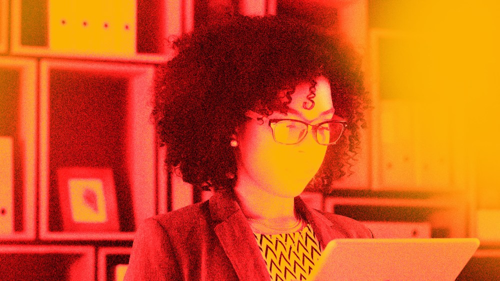 Photo illustration: Red and yellow overlay over an image of a bespectacled Black woman looking at a screen amidst file cabinets.