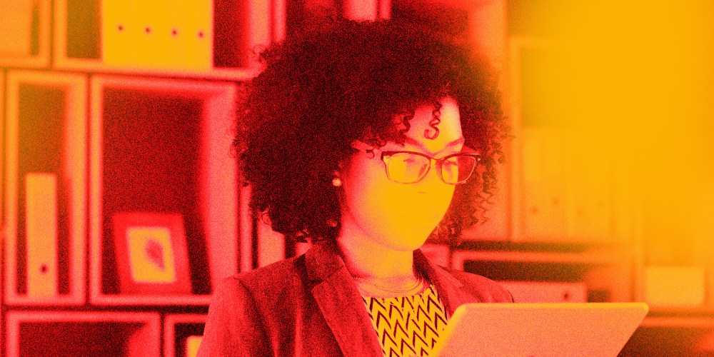 Photo illustration: Red and yellow overlay over an image of a bespectacled Black woman looking at a screen amidst file cabinets.
