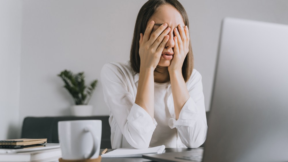 A woman sitting at desk holds her head in her hands.