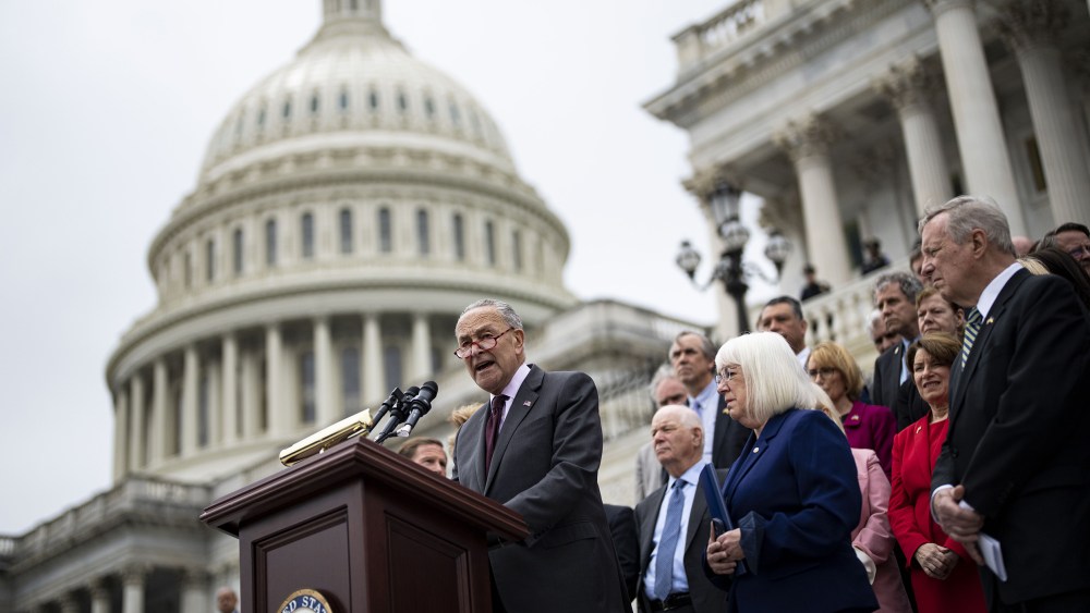 Image: Chuck Schumer speaks on the podium with the Senate Democrats standing behind him on the steps of the U.S. Capitol.