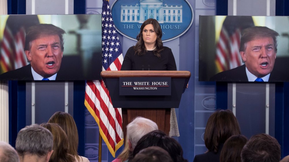 White House Press Secretary Sarah Huckabee Sanders stands beside monitors showing US President Donald J. Trump delivering a statement on the economy, at the beginning of a news conference in the James Brady Press Briefing Room of the White House, January