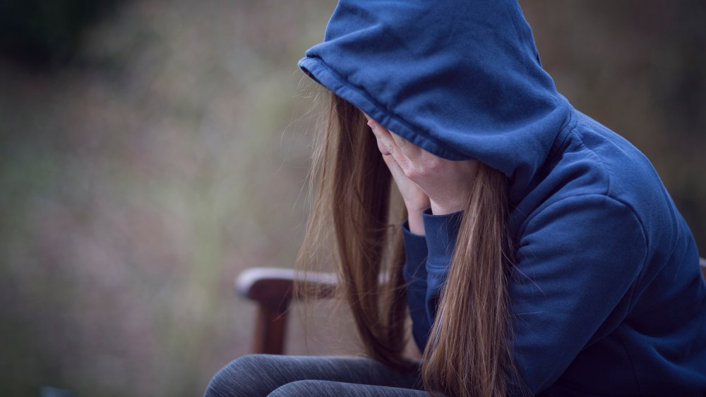 Image: Teenage girl in hooded top, with head in hands in despair