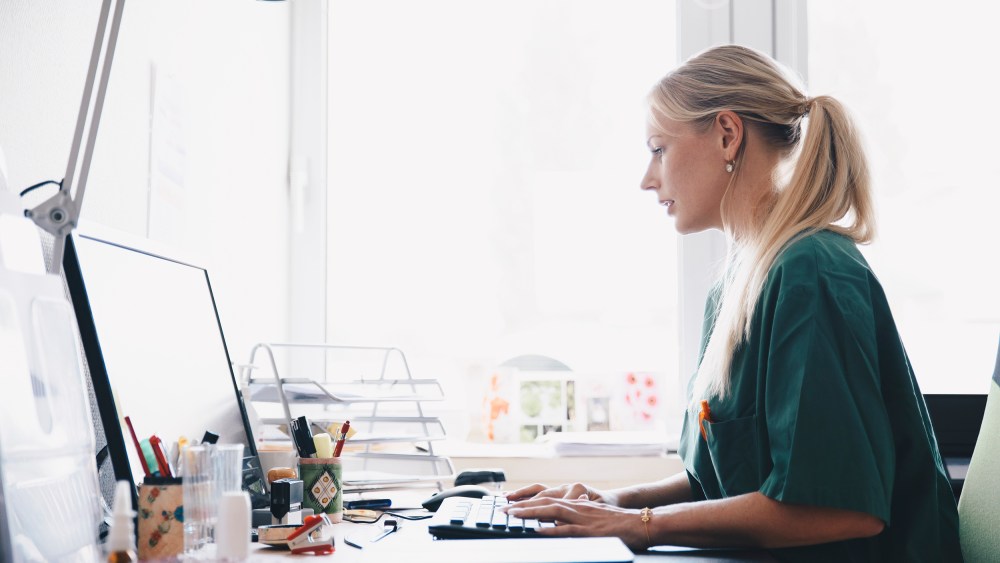 Image: Side view of female nurse working at computer desk against window in office