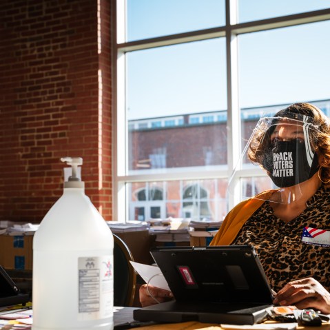 Image: An election official wearing a mask that reads "Black Lives Matter" sits at her desk speaking to a voter
