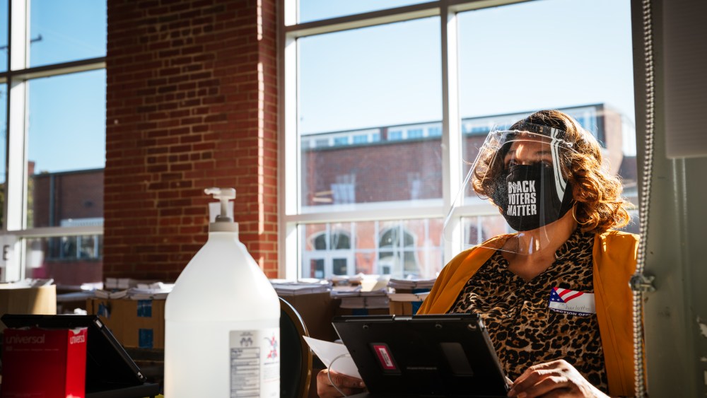 Image: An election official wearing a mask that reads "Black Lives Matter" sits at her desk speaking to a voter