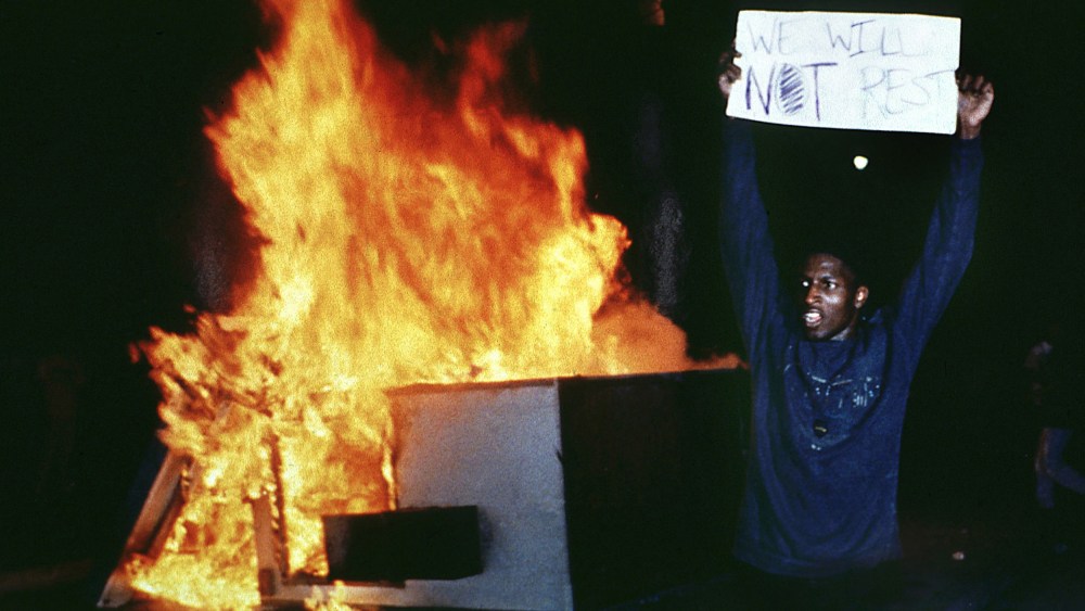 Image:A man holds up a sign that reads,"We will not rest" in front of a fire.