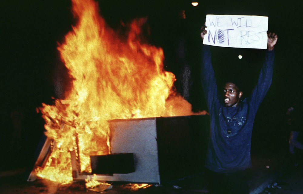 Image:A man holds up a sign that reads,"We will not rest" in front of a fire.