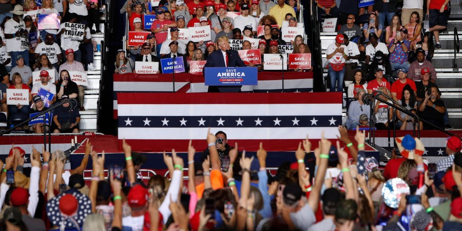 Image: Donald Trump on stage speaks to audience members who have their index fingers raised in unison.