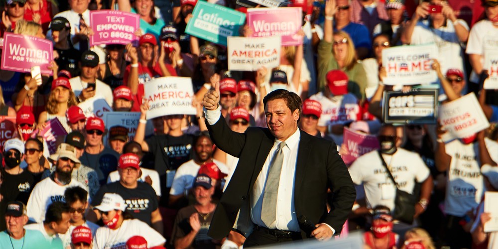 Image: Ron DeSantis speaks to a crowd during a campaign rally. A sign in the audience reads,"Latinos for Trump".
