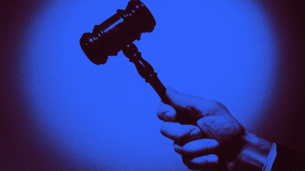 Image: A close up of a judge's hand holding a wooden gavel in a spotlight.