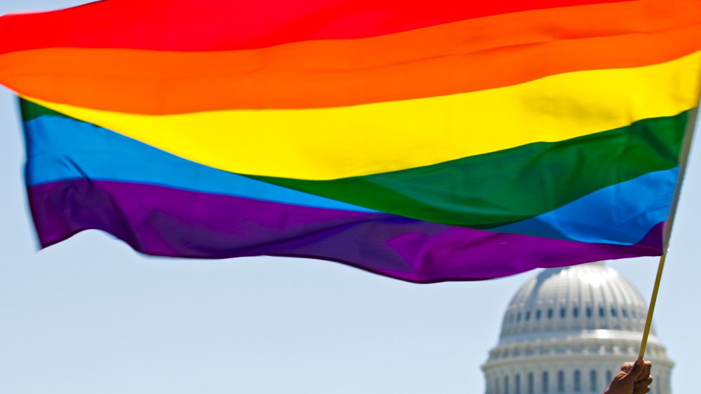 Image: A hand holds up a rainbow flag with the U.S. Capitol dome in the background.