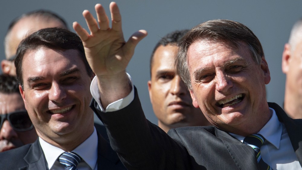 Image: Flavio stands next to Jair Bolsonaro who's waving during a ceremony.