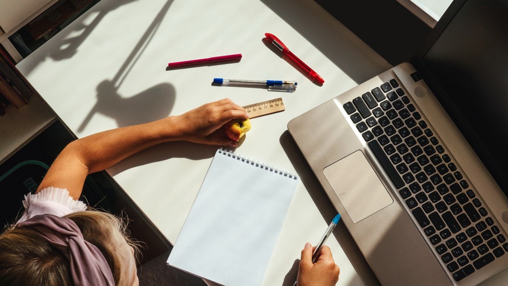A girl seated at a desk with a laptop, notebook and pens.