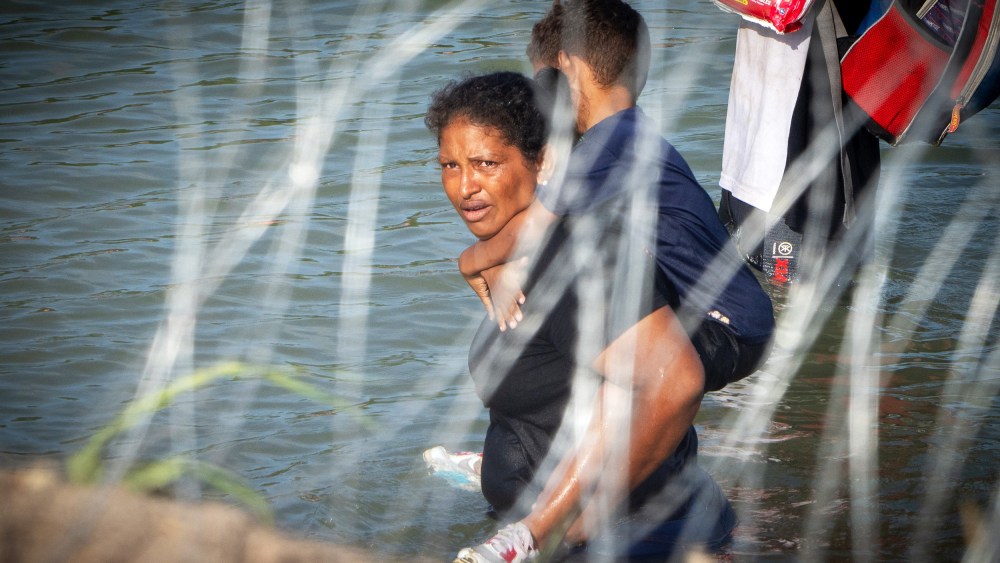 TOPSHOT - A woman holds a child on her back as migrants walk near concertina wire in the water along the Rio Grande border with Mexico in Eagle Pass, Texas, on July 16, 2023. A nearby buoy installation is part of an operation Texas is pursuing to secure its borders, but activists and some legislators say Governor Greg Abbott is exceeding his authority.