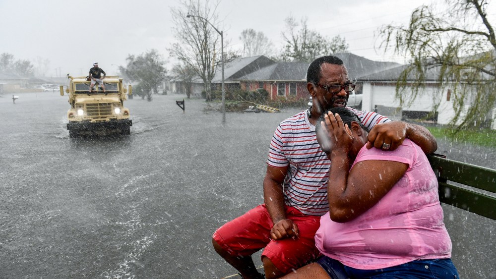 Two people embrace while sitting on a bench as a rain shower soaks them outside, as a volunteer evacuation truck is in the distance