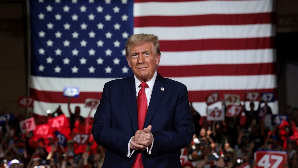 Donald Trump claps with an audience holding "47" signs behind him. A large American flag is the backdrop.