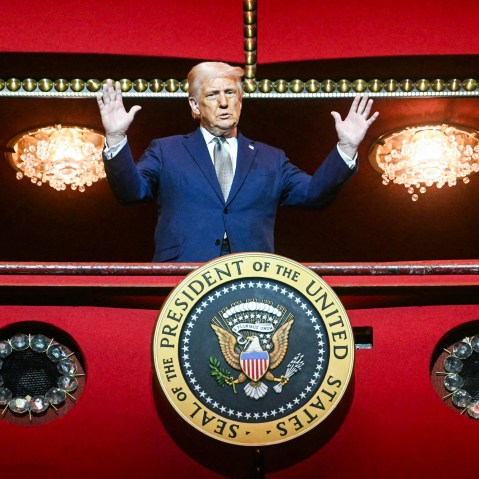Donald Trump, with both hands raised, stands on a balcony inside of a building