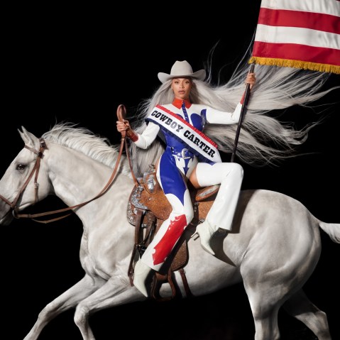 Beyonce sitting on a white horse wearing a sash that says "Cowboy Carter" and waving the American flag
