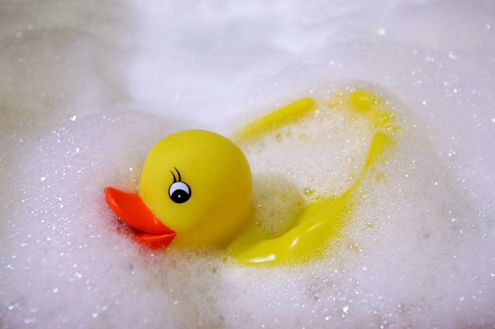 A rubber duck swims in a foam bath in a bath tub.