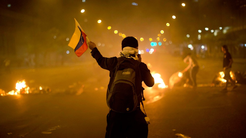 An anti-government demonstrator holds a representation of Venezuela's national flag in front of a burning barricade, Feb. 21, 2014, in the Altamira neighborhood of Caracas, Venezuela.