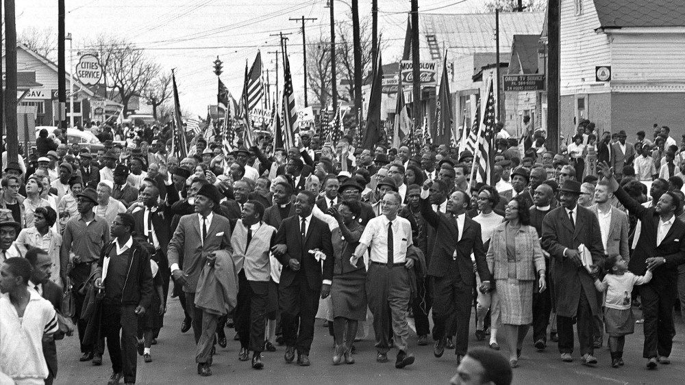 In this March 21, 1965 file photo, Dr. Martin Luther King, foreground row, fifth from right, waves as marchers stream across the Alabama River on the first of a five day, 50-mile march to the state capitol at Montgomery, Ala. (Photo by AP)
