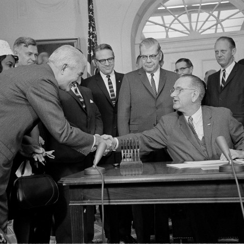 President Lyndon B. Johnson, who used 60 pens to sign a $1.2 billion college aid bill at the White House, hands one of the souvenirs to President Logan Wilson of the American Council on Education, Dec. 16, 1963. A room full of persons who helped pass...
