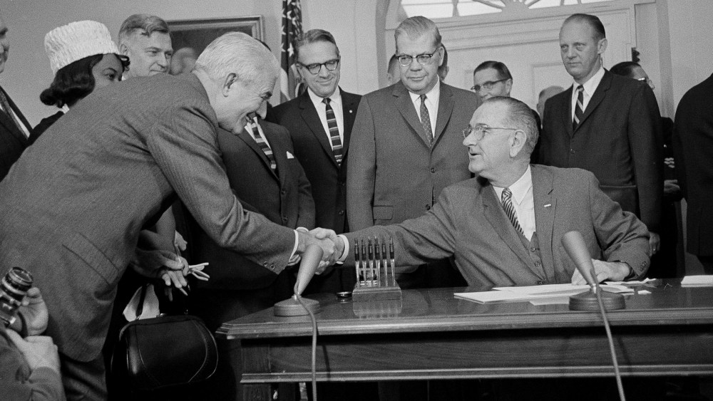 President Lyndon B. Johnson, who used 60 pens to sign a $1.2 billion college aid bill at the White House, hands one of the souvenirs to President Logan Wilson of the American Council on Education, Dec. 16, 1963. A room full of persons who helped pass...