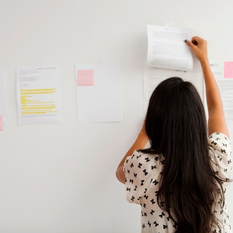A Businesswoman tapes up papers on an office wall. (Photo by Gallery Stock)