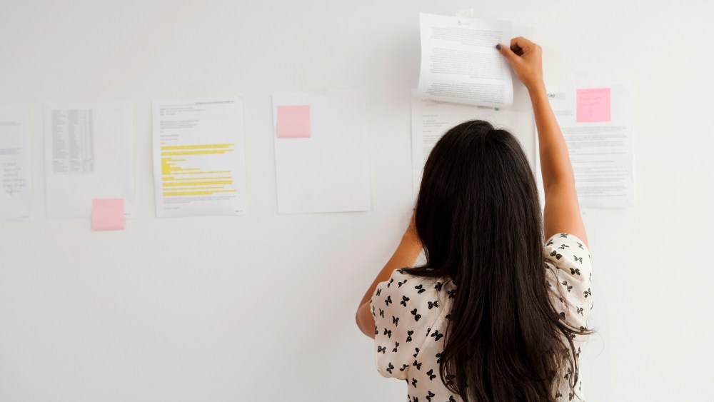 A Businesswoman tapes up papers on an office wall. (Photo by Gallery Stock)