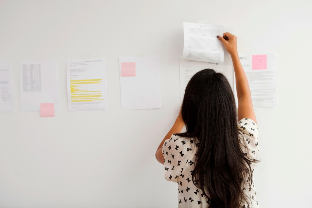 A Businesswoman tapes up papers on an office wall. (Photo by Gallery Stock)