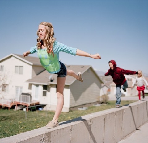 A young teeanage girl does gymnastics moves while balancing on a concrete ledge as her little brother and sister copy her with a view of mountains and her family's suburban neighborhood in background. Pleasant Grove, Utah. Brian Shumway/Redux