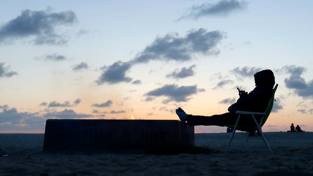 A woman sits by an open fire at Moonlight Beach as she reads messages on her phone after sunset in Encinitas, California, March 31, 2014.